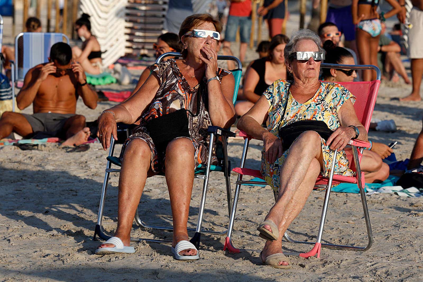 Two women wearing solar eclipse glasses sit in beach chairs on a sandy beach with other sunbathers