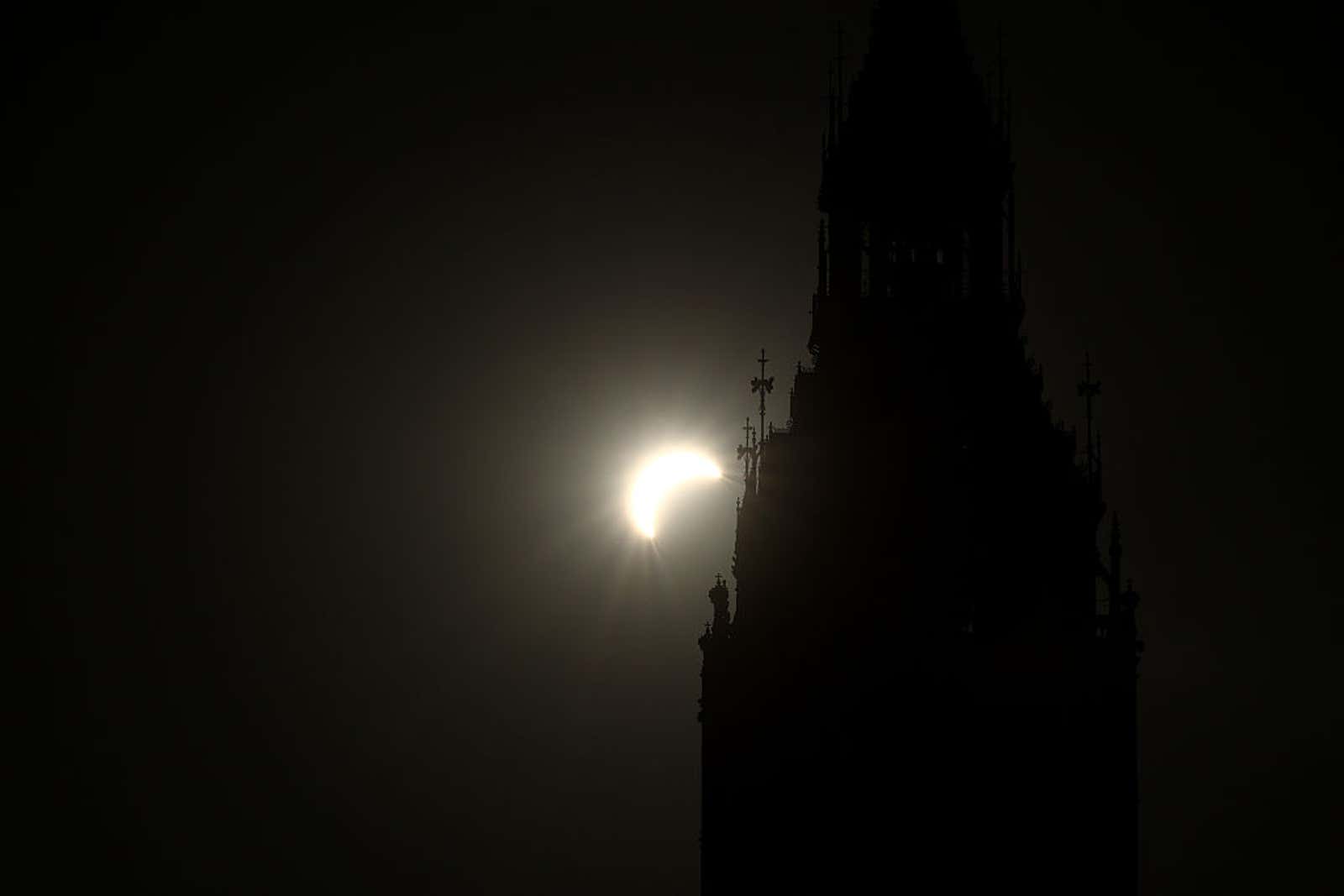 Crescent-shaped partial solar eclipse glowing beside a dark silhouetted Gothic clock tower spire