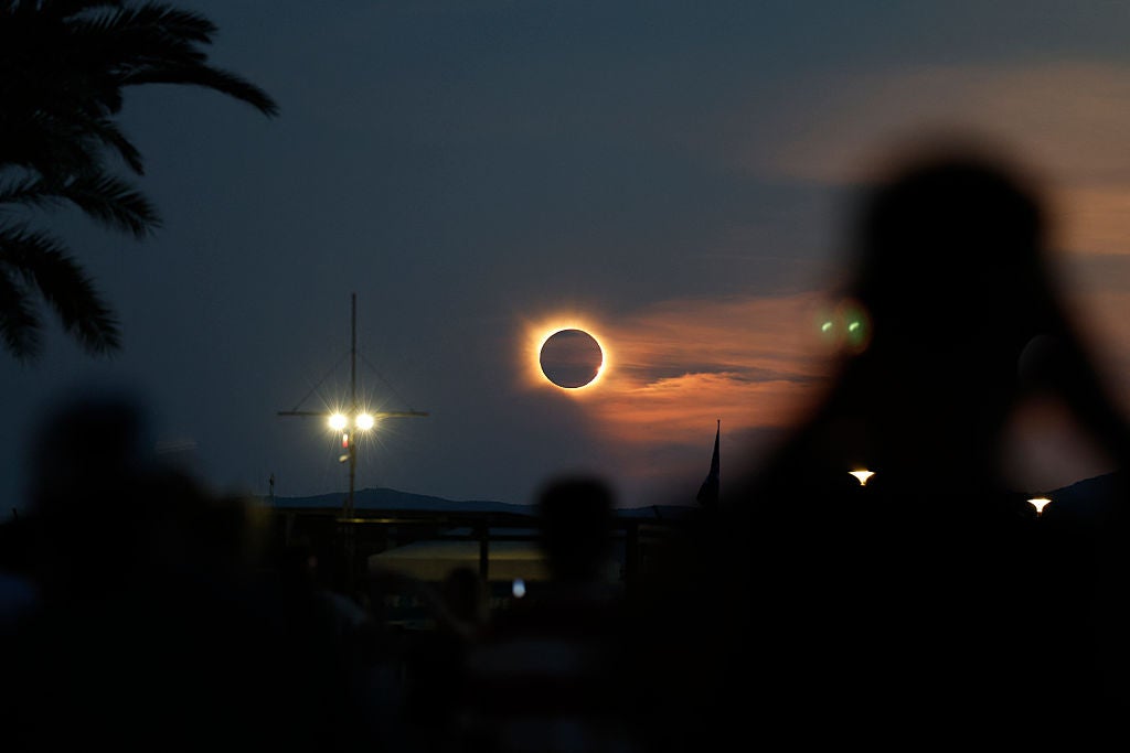 Solar eclipse showing bright ring of light around dark moon at twilight, with silhouetted onlookers and palm tree in foreground