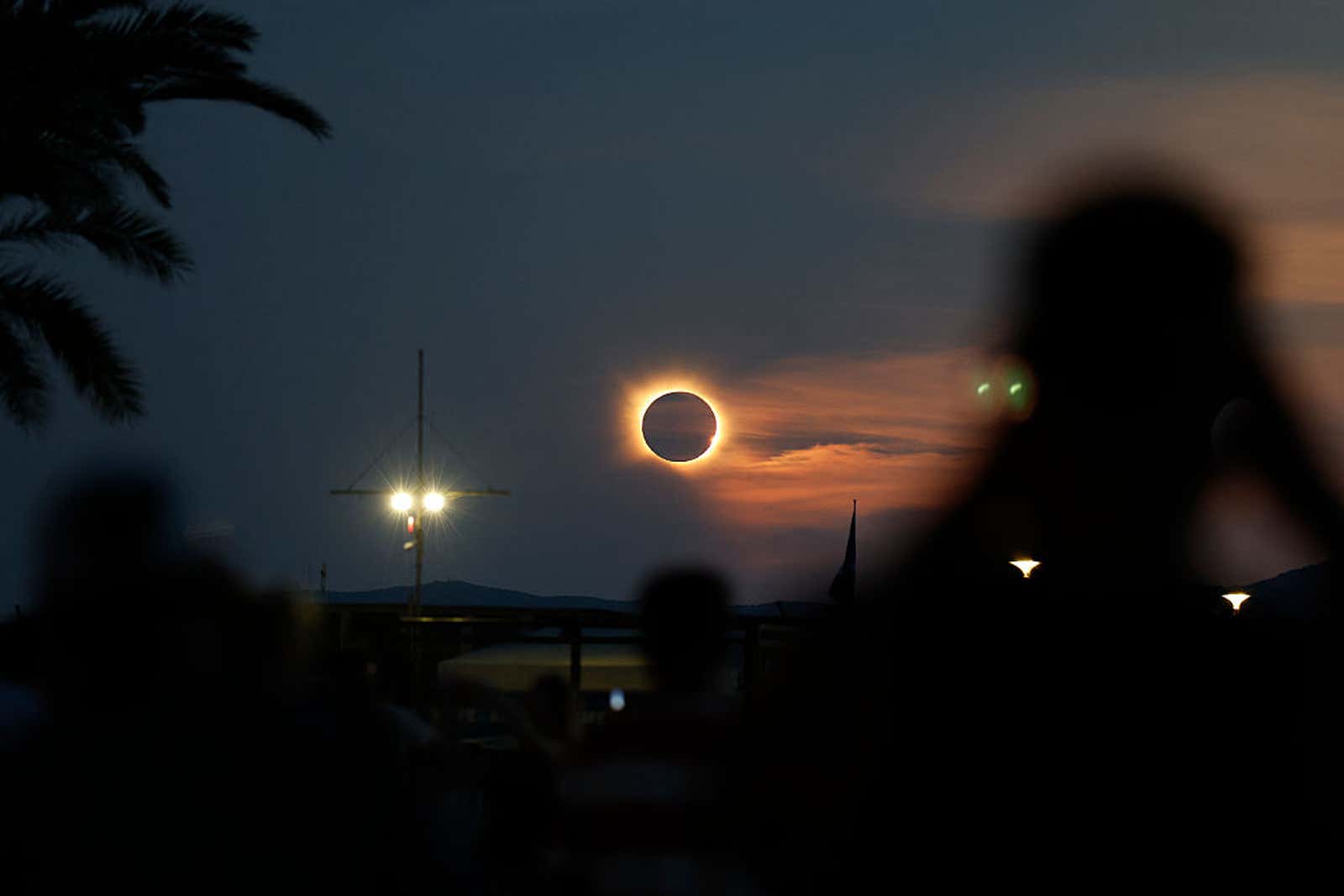 Solar eclipse showing bright ring of light around dark moon at twilight, with silhouetted onlookers and palm tree in foreground