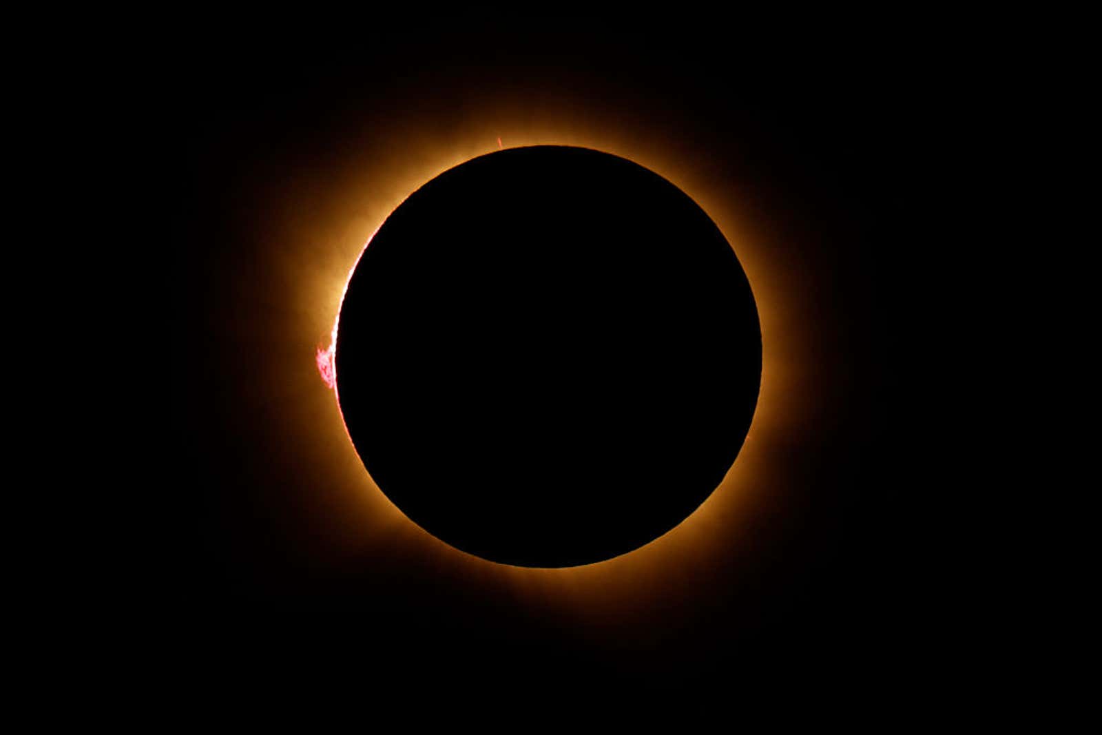 Total solar eclipse showing dark moon silhouette surrounded by glowing golden corona and a red solar prominence