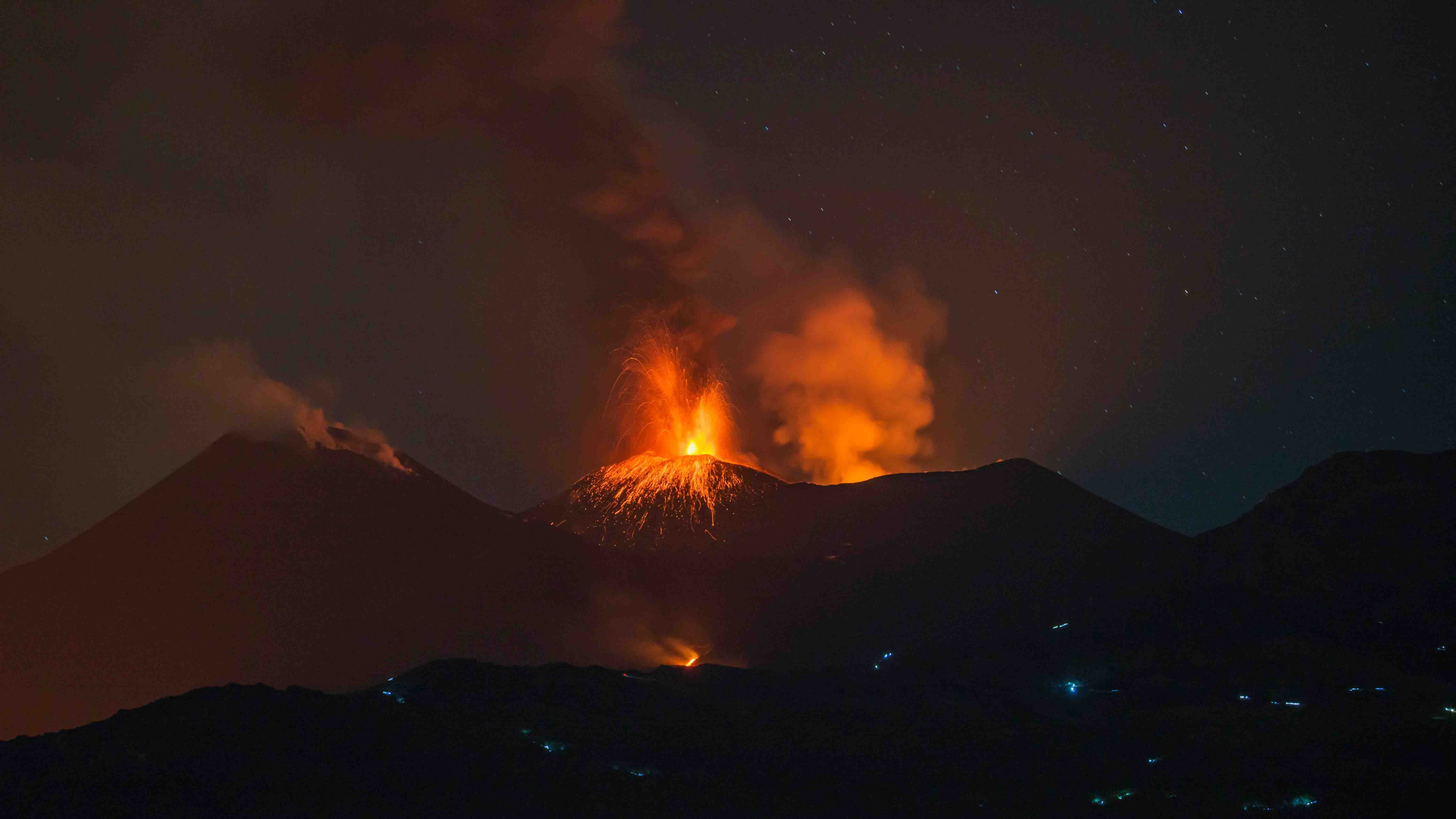 Volcano erupting at night with glowing lava spewing from the crater and orange smoke rising against a starry sky