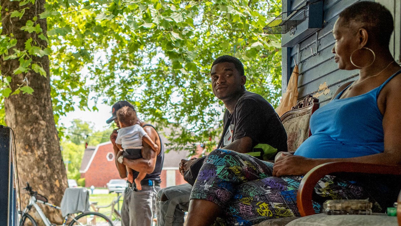 Family relaxing on porch steps under leafy tree, with man in dark shirt, woman in blue tank top, and person holding child in background