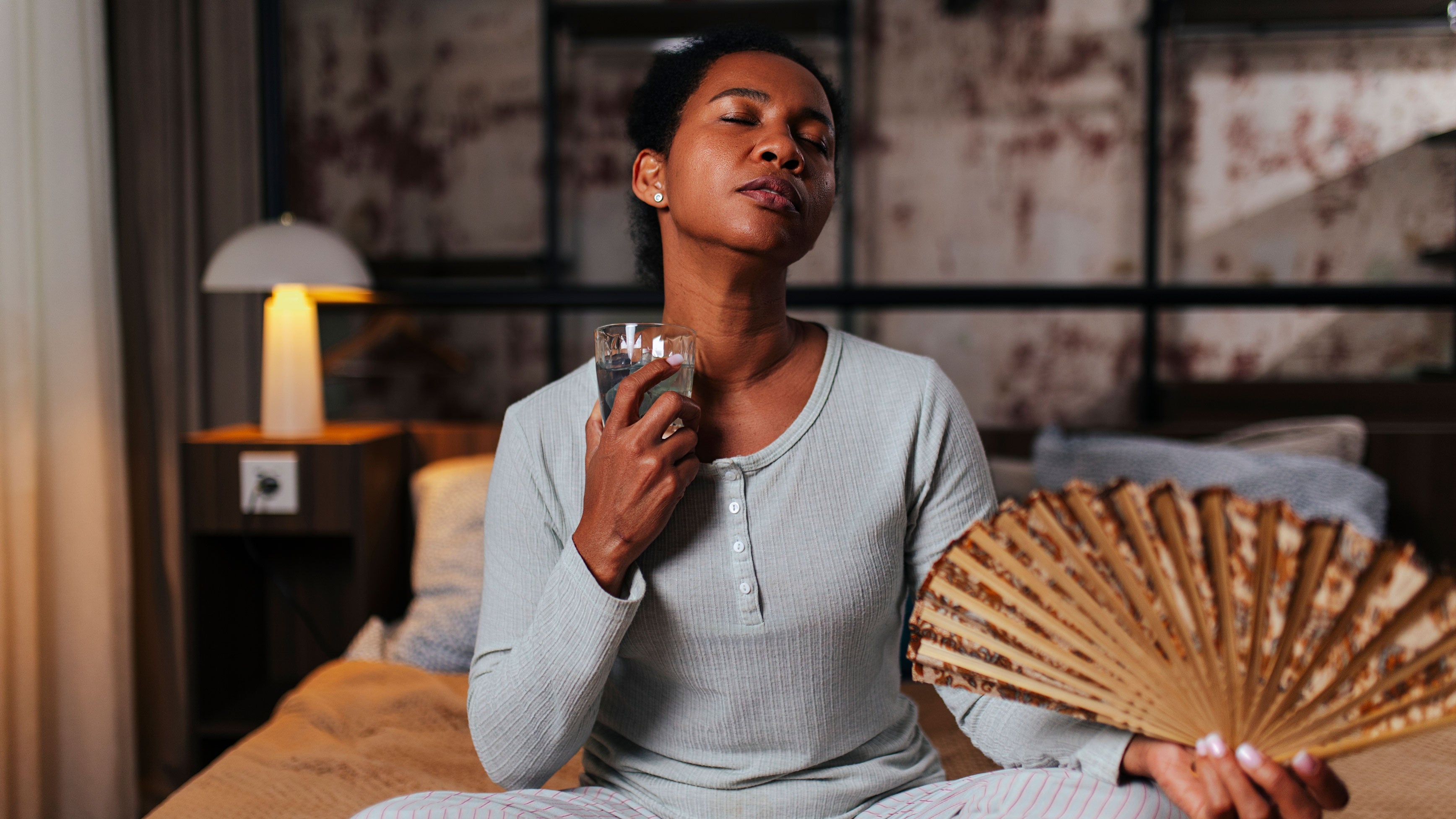 A woman in pajamas sitting on bed, drinking ice water and fanning herself during heat wave.