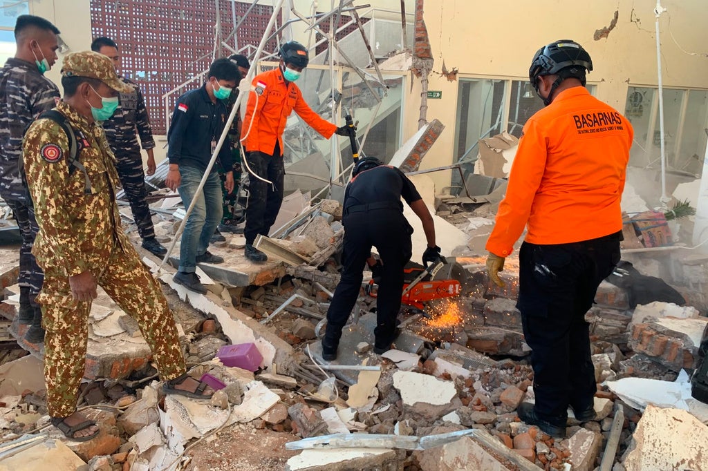 BASARNAS rescue workers and military personnel search through earthquake rubble at a collapsed building, using a cutting tool to clear debris