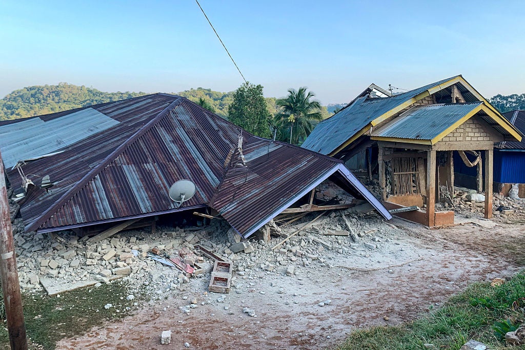 Collapsed house with rusted metal roof lying in rubble beside a partially damaged building after a disaster