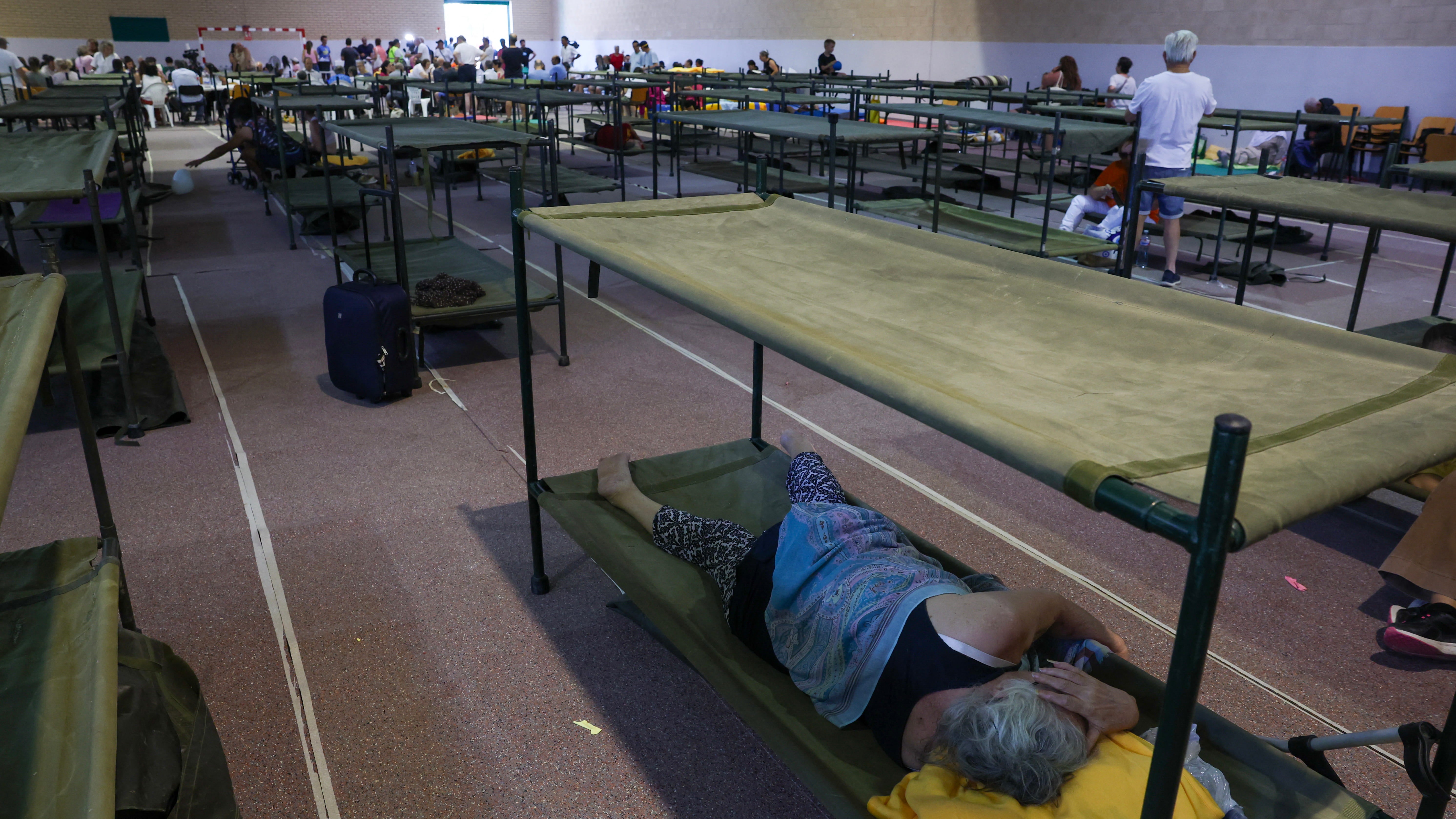 People resting on cot beds in a large indoor shelter or evacuation hall