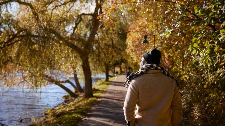 Person wearing a beanie and scarf walking along a tree-lined riverside path in autumn.