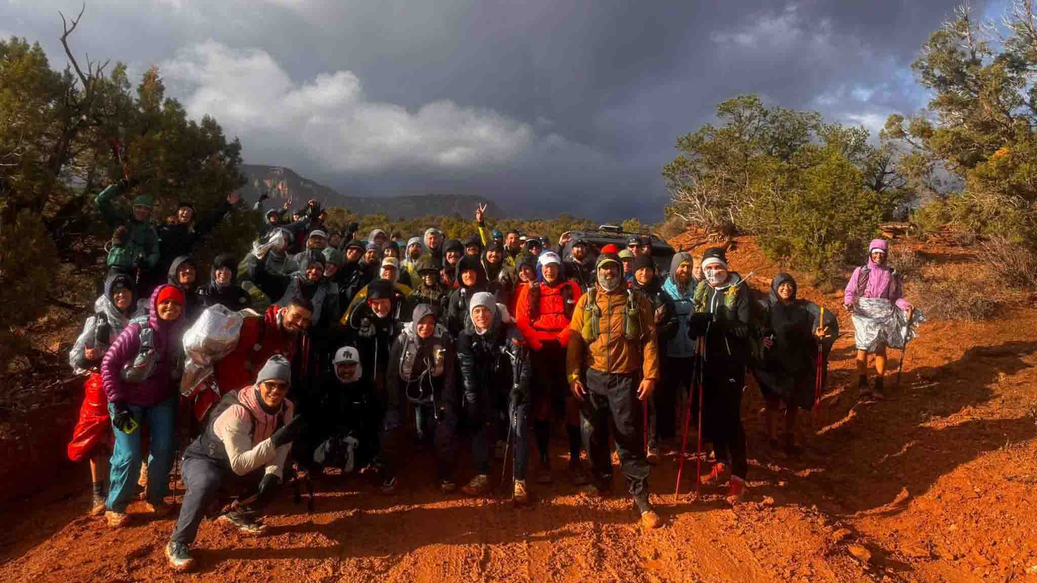 Large group of hikers in cold-weather gear posing on red dirt trail under dramatic cloudy sky