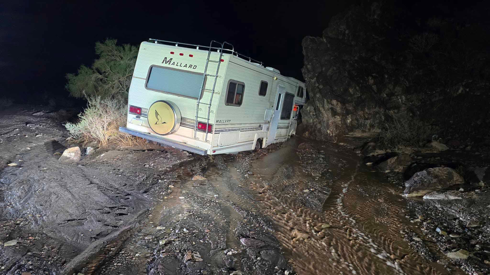A Mallard RV stuck between rocks in a muddy, flooded desert wash at night, illuminated by a spotlight.