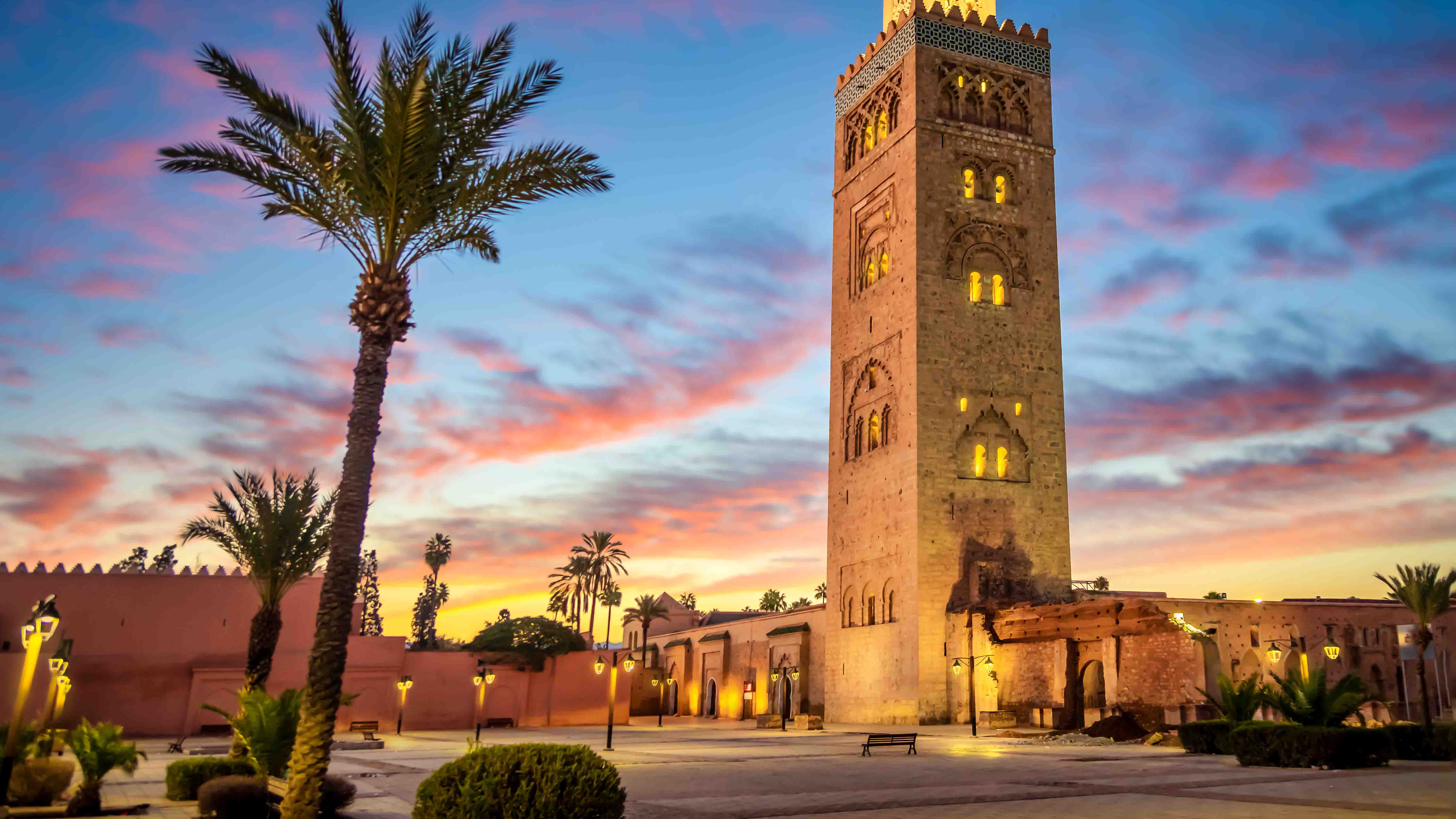 Illuminated Koutoubia Mosque minaret in Marrakech at sunset with palm trees and colorful sky