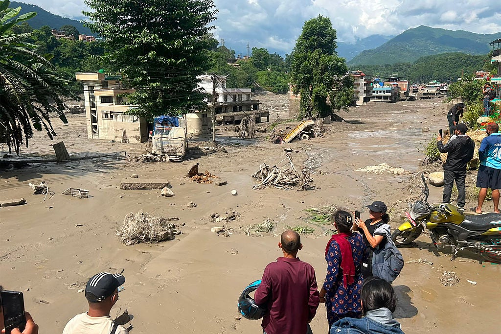 People watch and photograph a flood-devastated area with mud-covered debris, damaged buildings, and mountains in the background