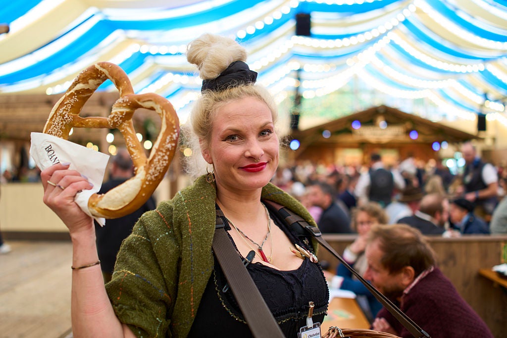 Waitress in traditional dress smiling while holding a large pretzel inside a crowded Oktoberfest beer tent.