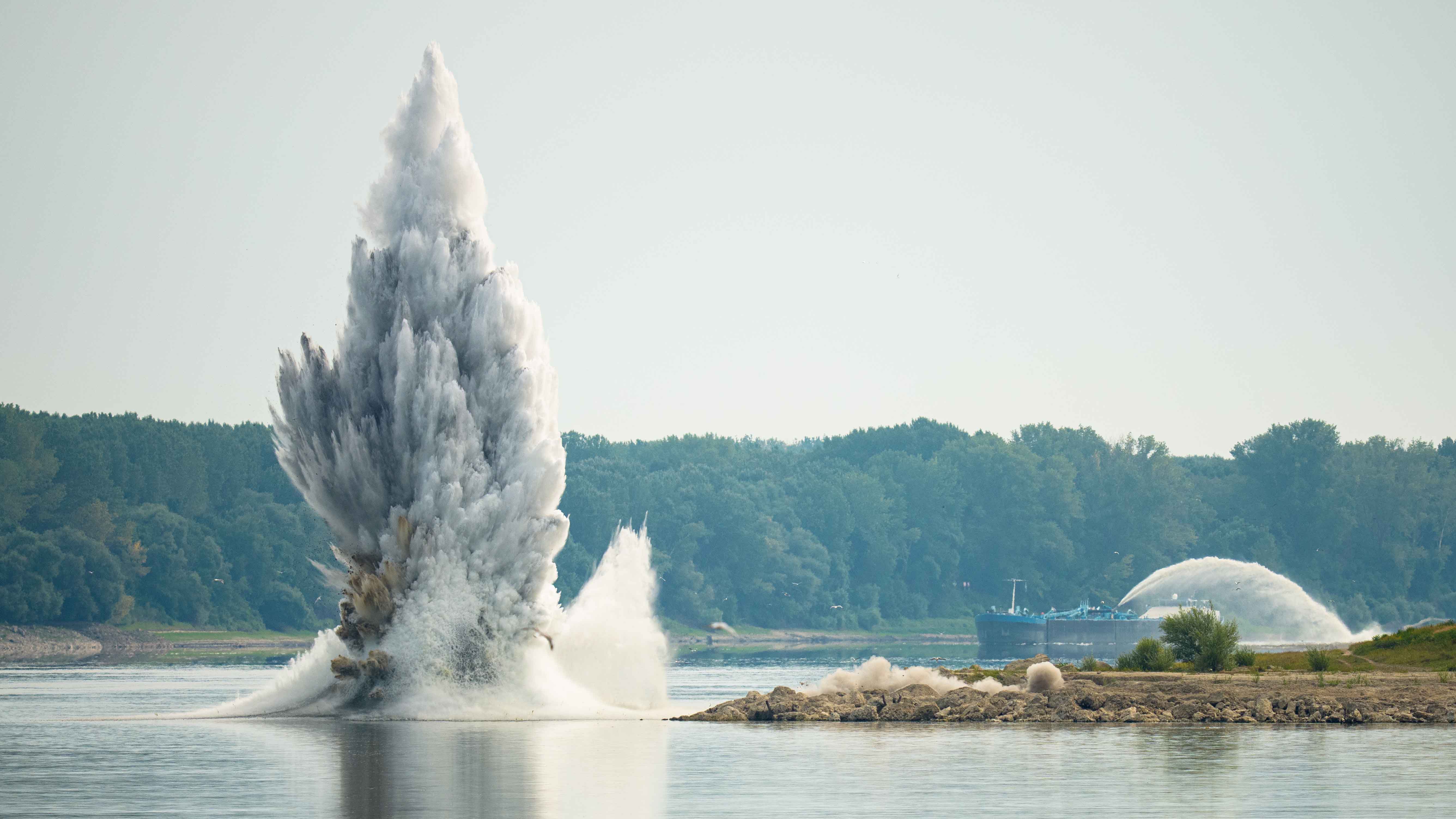 Large water explosion plume erupting from a river near a sandy bank with a barge and forested shoreline in the background