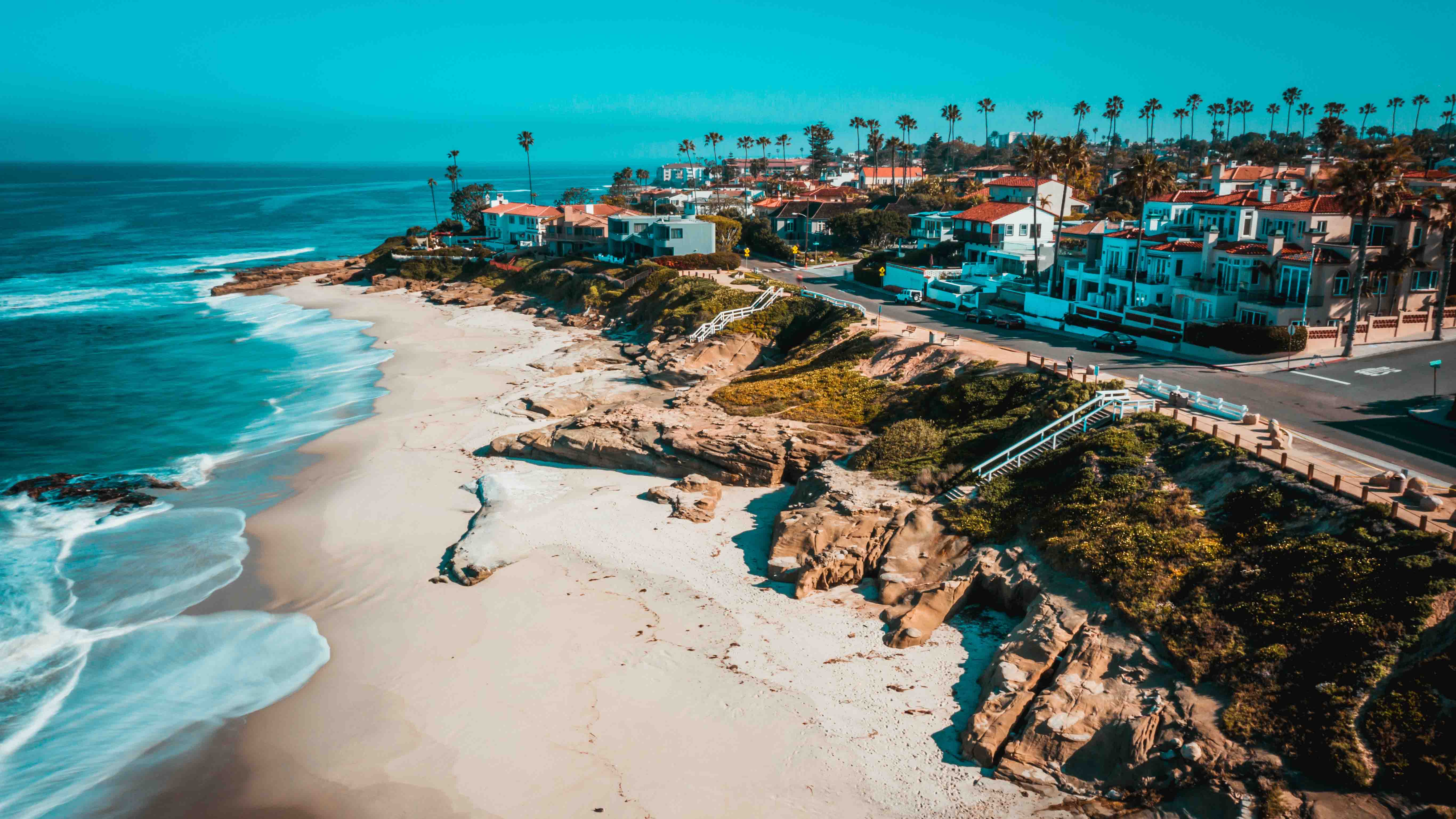 Aerial view of a coastal neighborhood with palm trees, beachfront homes, rocky cliffs, and turquoise ocean waves