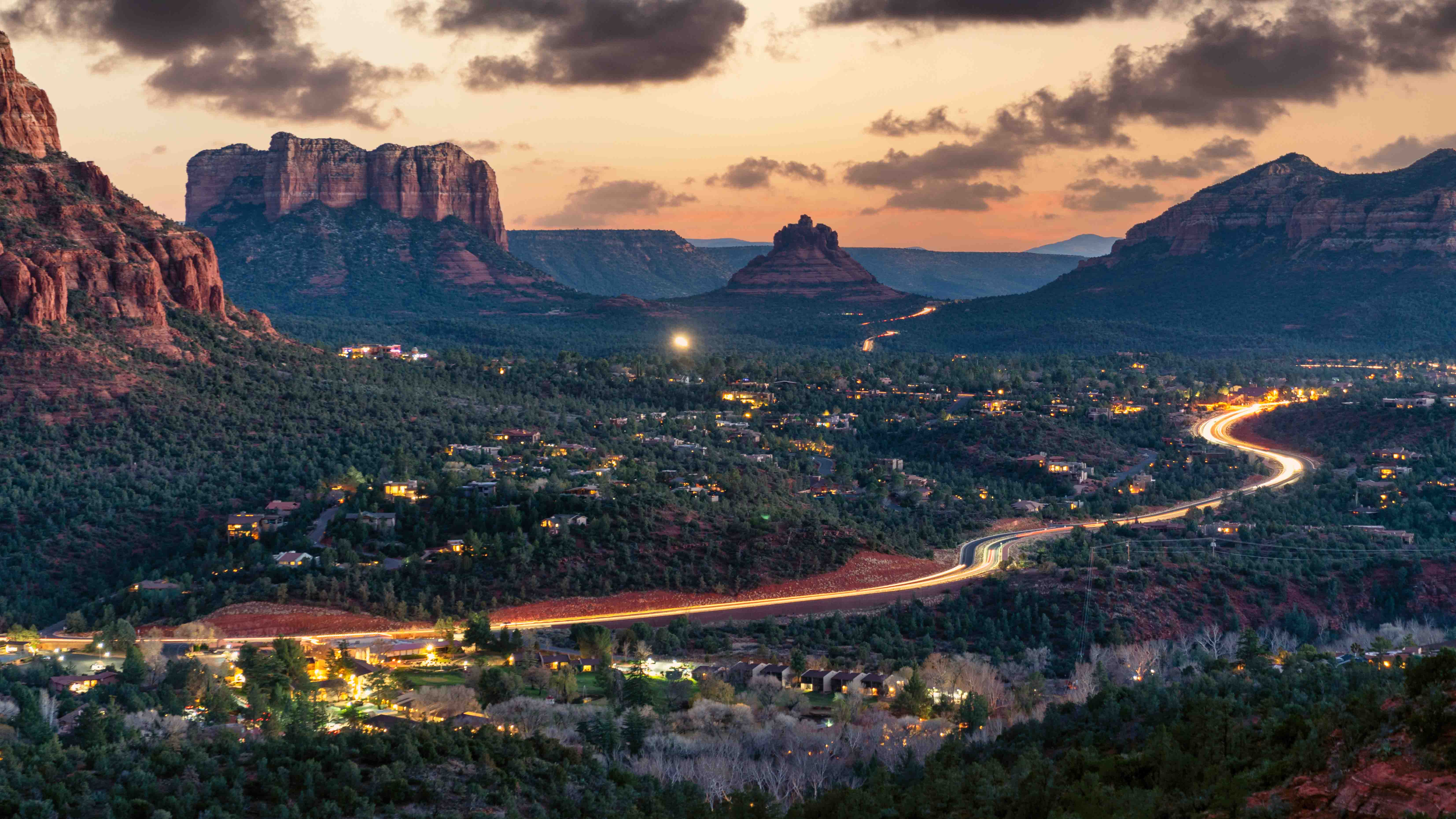 Aerial twilight view of Sedona, Arizona with red rock formations, a winding illuminated highway, and city lights below