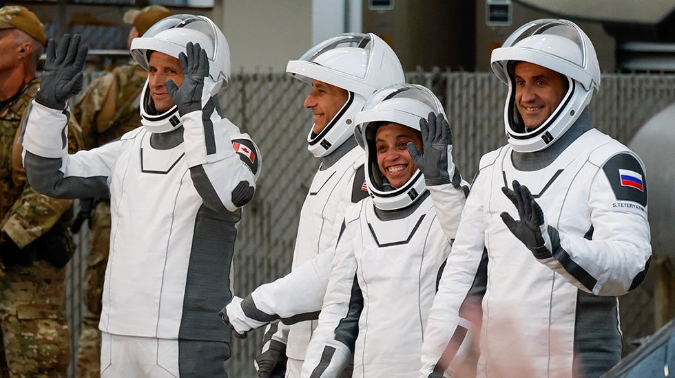 Four astronauts in space suits wave and smile