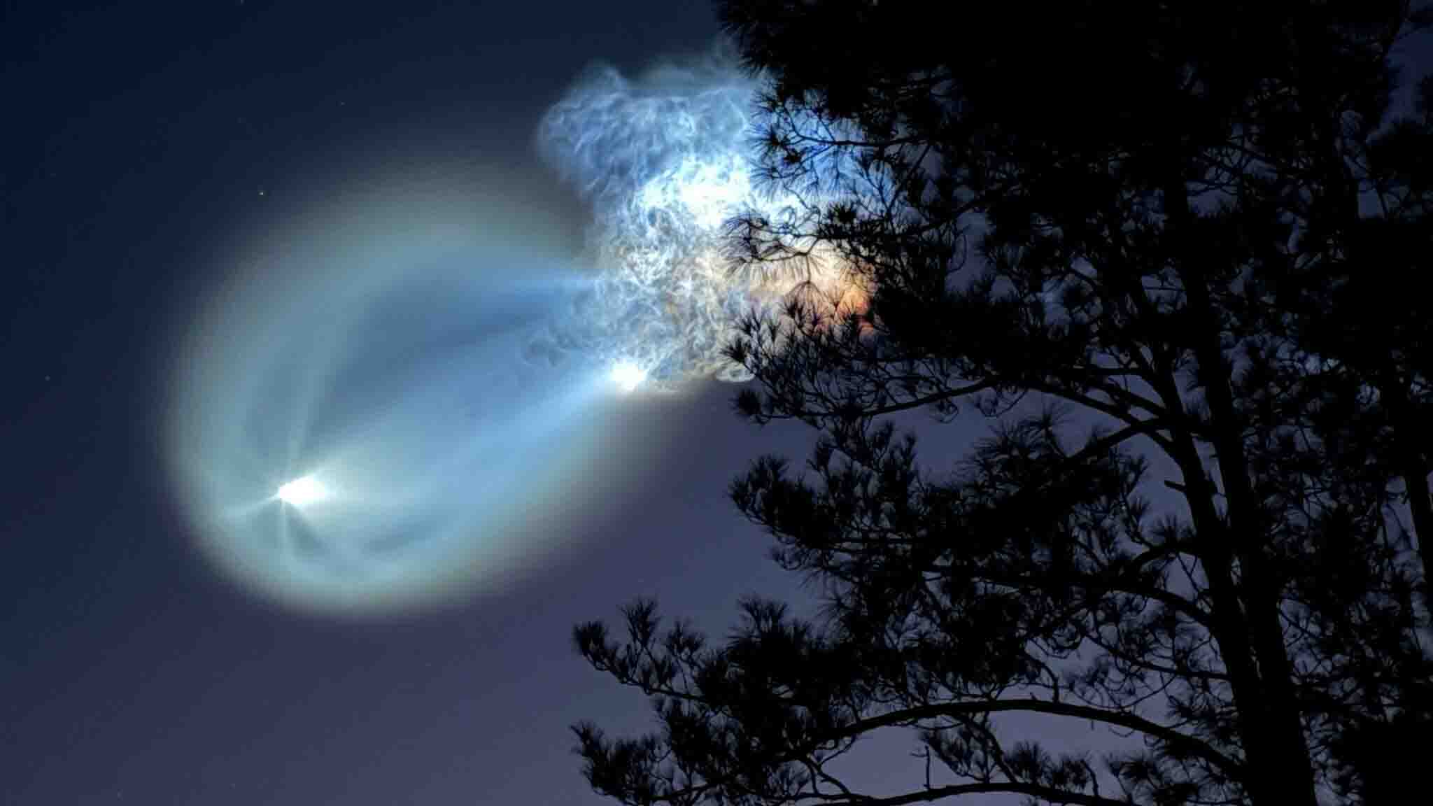 Glowing rocket exhaust plume illuminates the night sky behind silhouetted pine tree branches