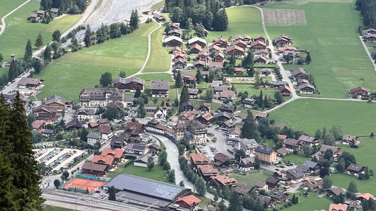 Aerial view of a Swiss alpine village with chalet-style buildings, a river, church, and surrounding green fields