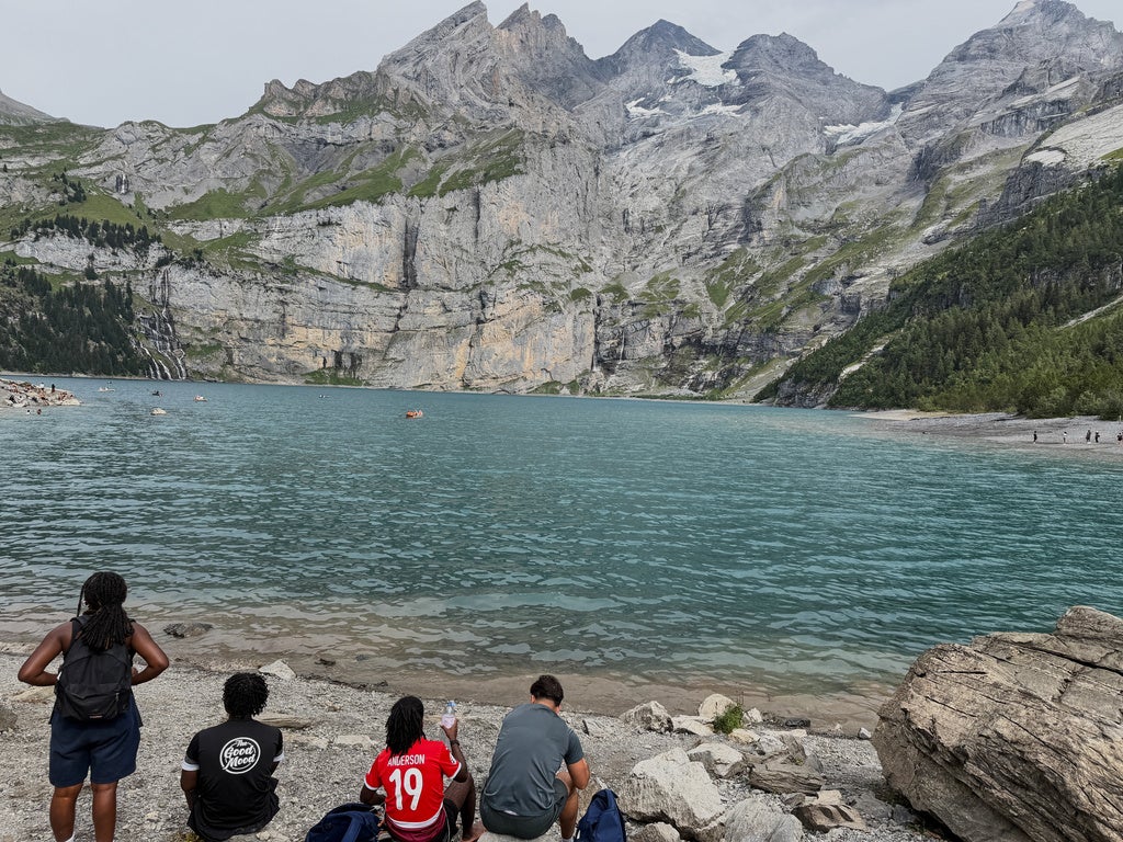 People sitting on rocky shore of turquoise alpine lake surrounded by towering gray mountains, likely Oeschinen Lake in Switzerland
