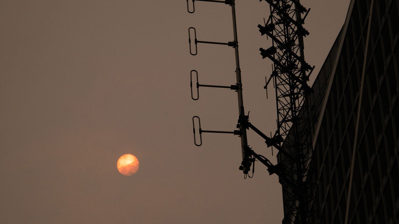 Orange sun in hazy sky beside silhouetted radio tower and building facade