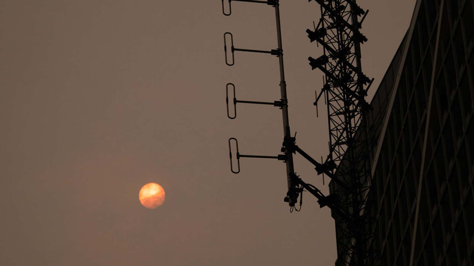 Orange sun in hazy sky beside silhouetted radio tower and building facade