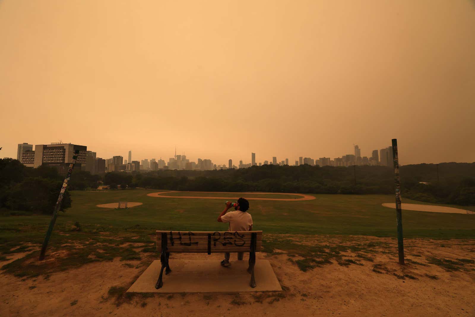 Person sitting on a graffiti-covered bench overlooking a park and city skyline under an orange sky