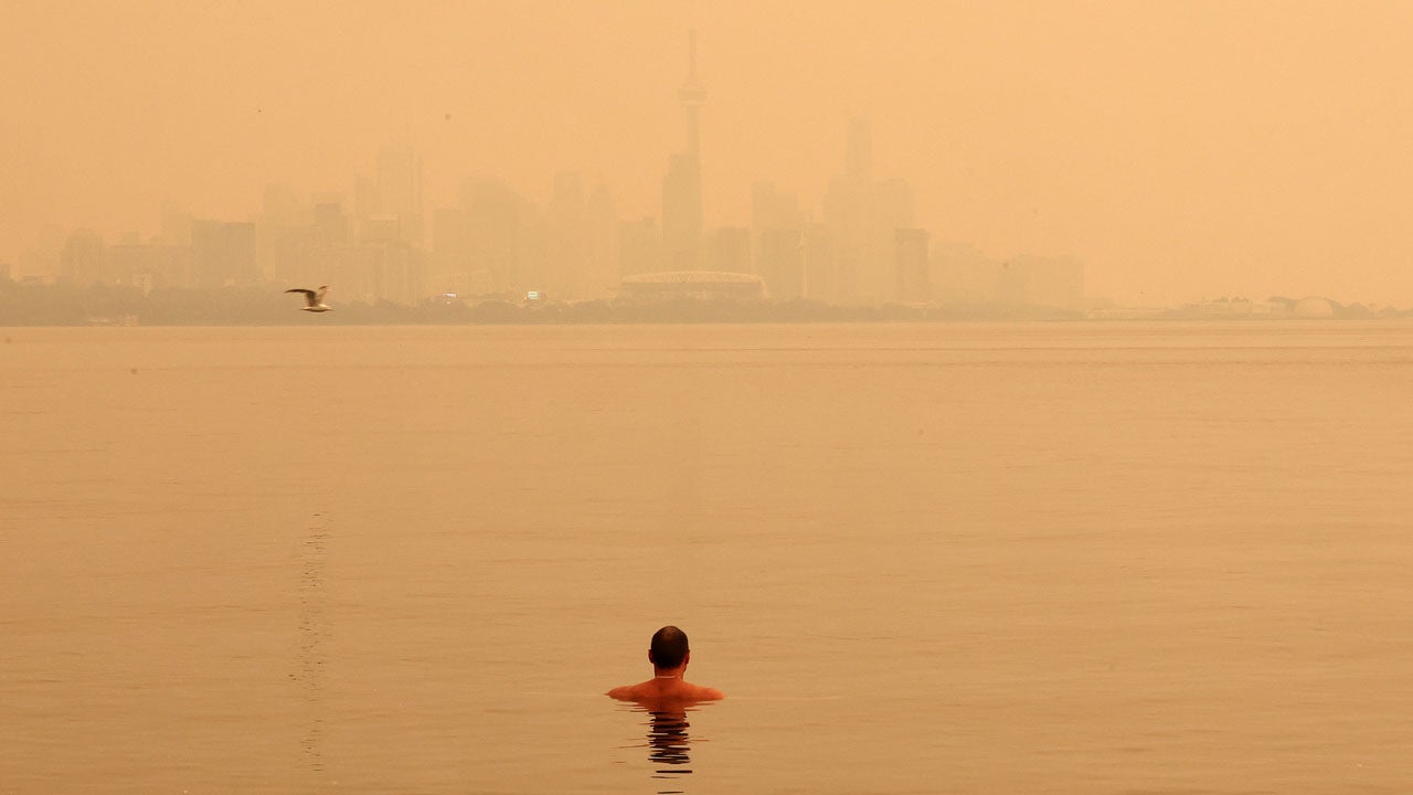 A lone swimmer in calm water faces a hazy city skyline under an orange sky