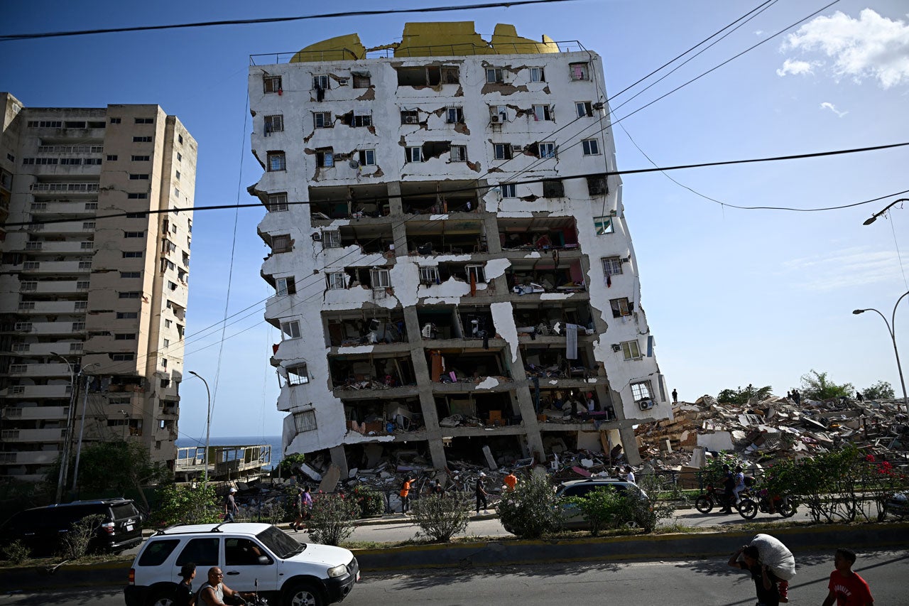Severely damaged white apartment building with exposed floors and missing walls, surrounded by debris and intact buildings