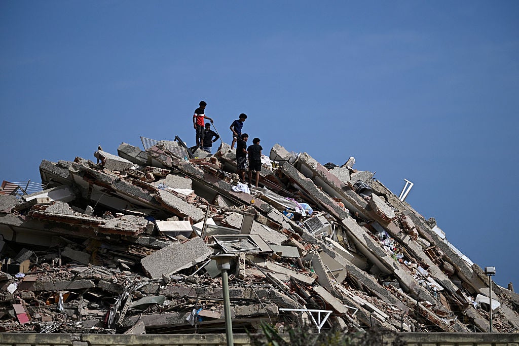 Several people stand on top of a large pile of concrete rubble and debris from a collapsed building against a clear blue sky