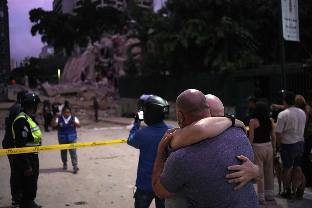 Two men embrace in foreground while emergency responders and crowds gather near collapsed building debris with yellow caution tape