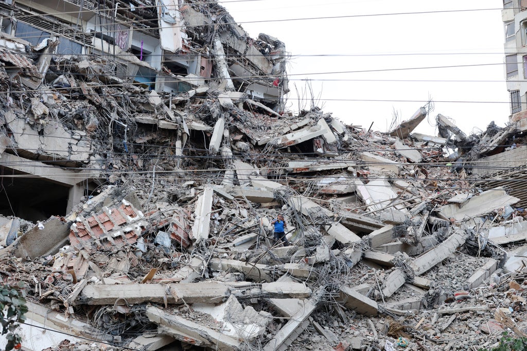 Collapsed multi-story building with concrete debris, twisted rebar, and two people in the rubble pile