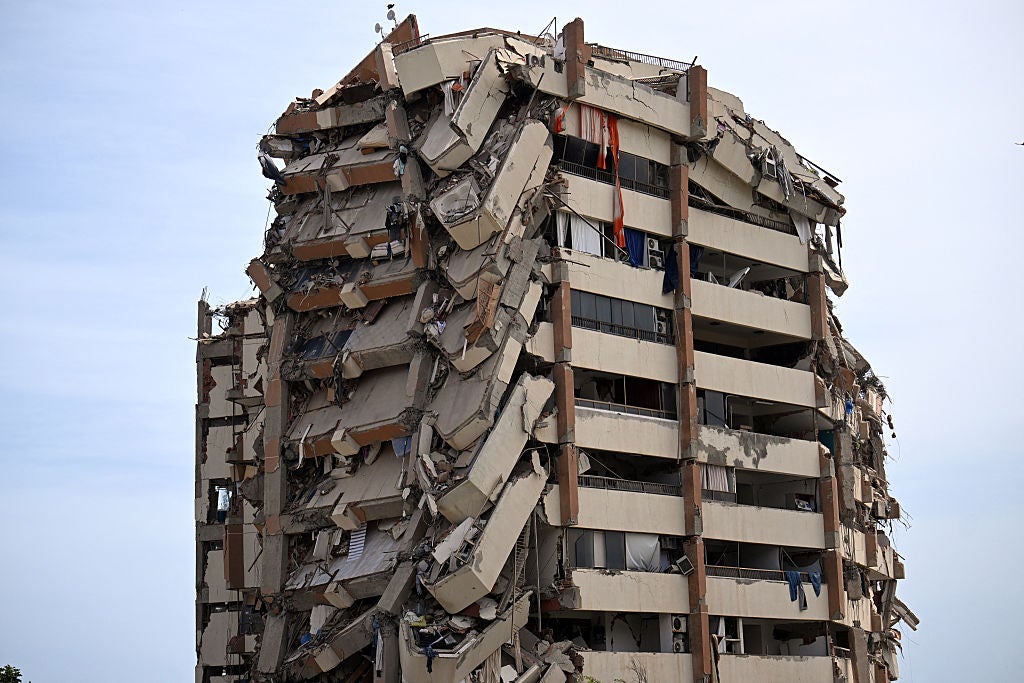 Severely damaged multi-story apartment building with collapsed sections, exposed concrete slabs, and debris hanging from the structure against a cloudy sky