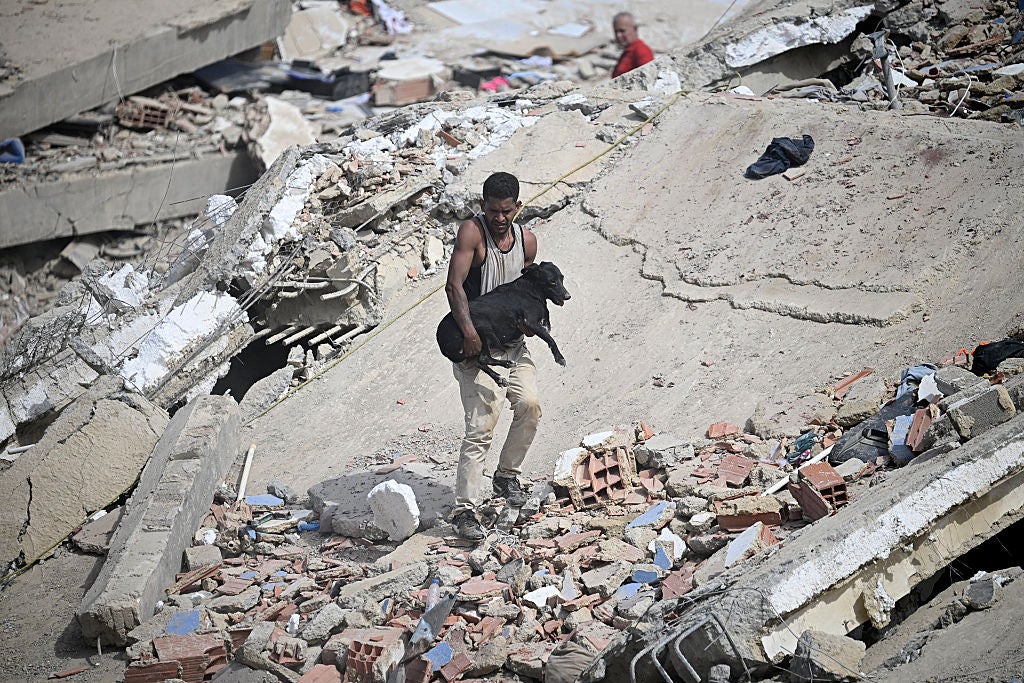 A man carrying items walks through extensive building rubble and debris scattered across broken concrete slabs