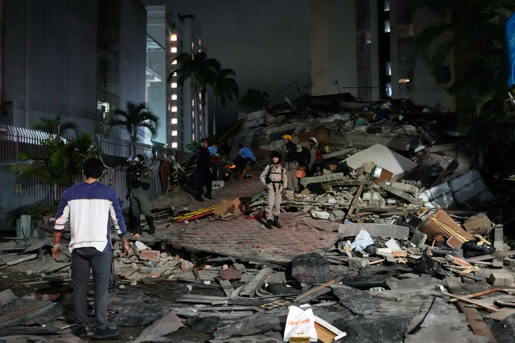 Rescue workers search through collapsed building debris at night with apartment buildings and palm trees visible in the background