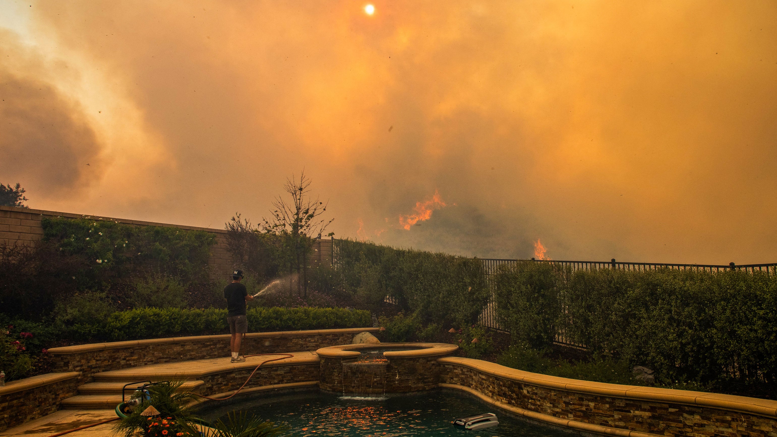 A resident uses a garden hose to watering his backyard as the Sandy Fire burns in the hills close to a residential area in Simi Valley, California, on May 18, 2026. About 24,000 people in Simi Valley, 40 miles northwest of Los Angeles, are under mandatory evacuation orders as the Sandy Fire is "spreading dangerously fast" according to fire officials.