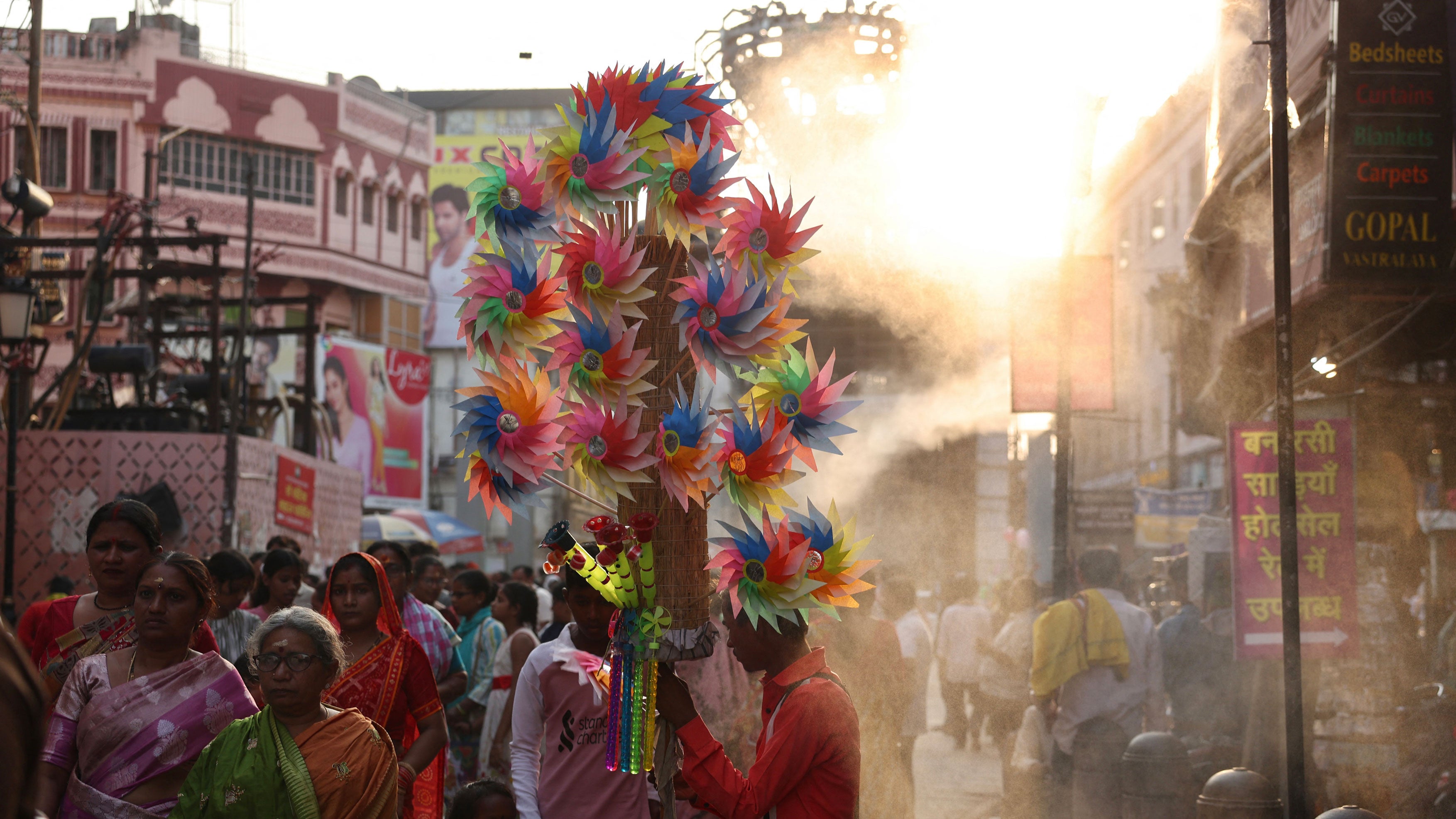 People walk under water sprinklers installed along a street on a hot summer day in Varanasi on May 15, 2026.