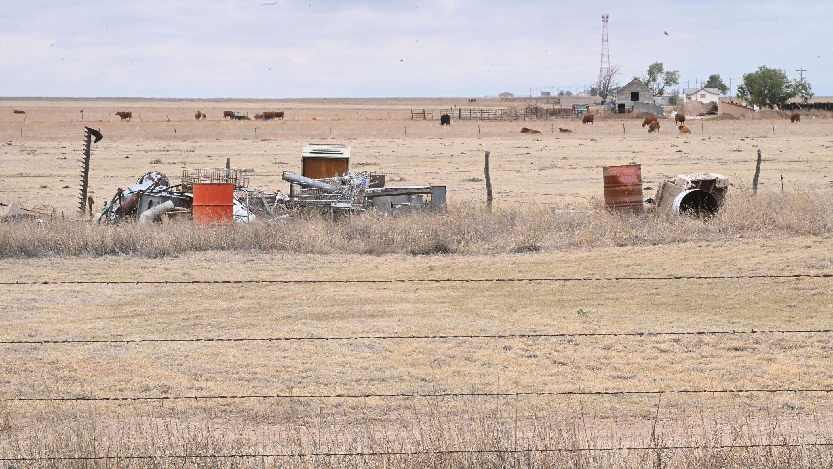 Rural farmyard with scattered equipment and debris in foreground, cattle grazing in fenced pastures, and farm buildings in the distance under cloudy sky
