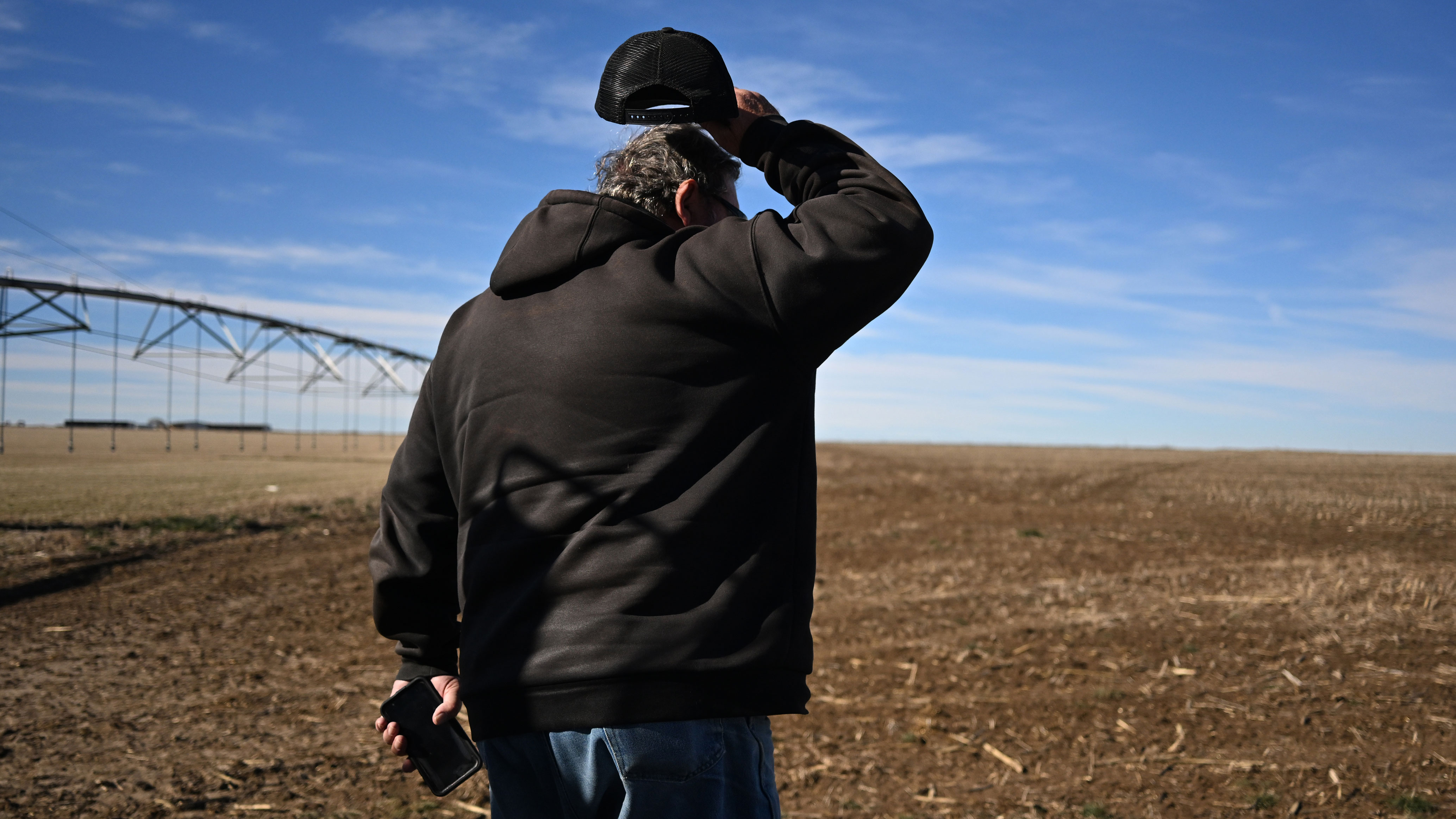 Man in black hoodie and cap looking across a brown agricultural field with irrigation equipment visible
