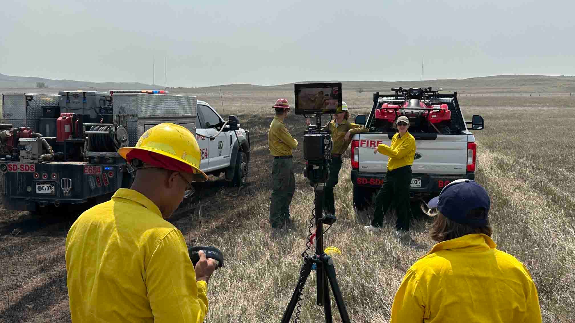 Wildland firefighters in yellow gear filming a training video near fire trucks in a grassy field