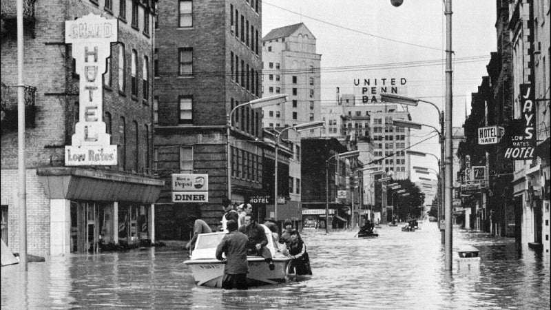 Black and white photo of people in a boat navigating a flooded city street lined with hotel and diner signs